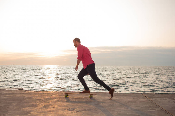 skater in red shirt and blue jeans riding near beach on longboard during sunrise, sea or ocean background - Foto, imagen