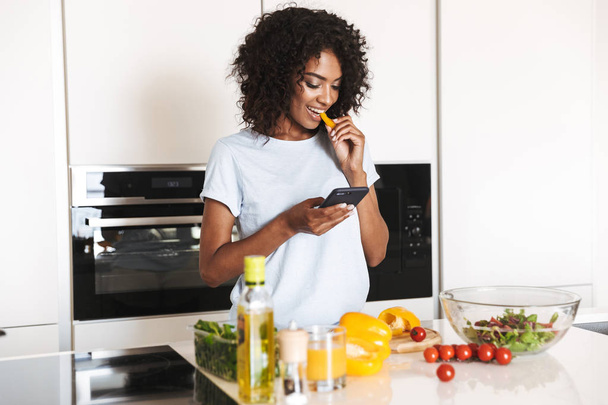 Portrait of a happy afro american woman using mobile phone while making a healthy salad at the kitchen - 写真・画像