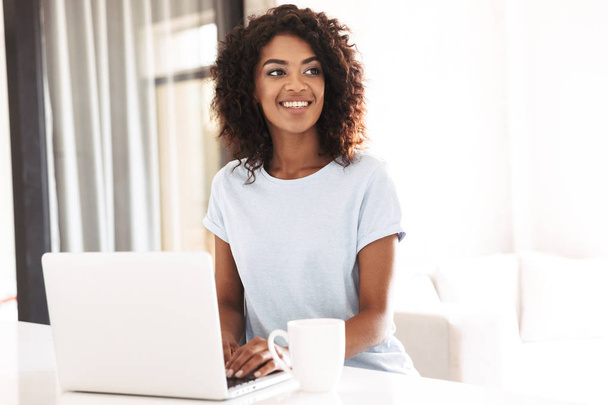 Cheerful african woman using laptop computer while sitting at home with cup of coffee - Photo, image