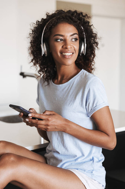 Attractive african woman in headphones using mobile phone while sitting on a chair at home - Foto, immagini