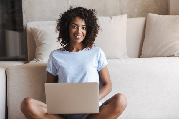 Vidám afrikai nő használ laptop számítógép, miközben ül a kanapén, otthon - Fotó, kép