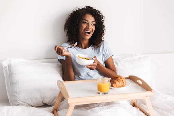 Laughing young african woman having tasty breakfast on a tray in bed - Фото, изображение