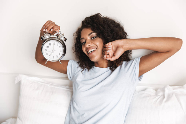 Smiling young african woman showing alarm clock while sitting in bed in the morning - Zdjęcie, obraz