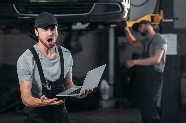 shocked mechanic using laptop, while colleague working in workshop behind - Photo, Image