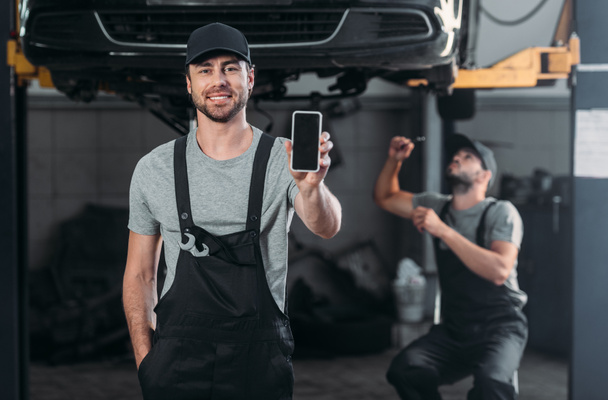 auto mechanic presenting smartphone with blank screen, while colleague working in workshop behind - Photo, Image