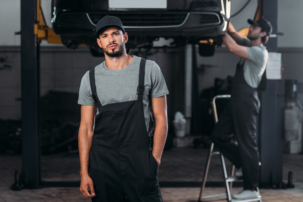 auto mechanic posing in overalls, while colleague working in workshop behind - Photo, Image