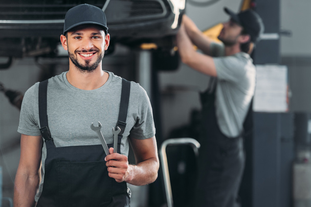 smiling auto mechanic holding wrenches, while colleague working in workshop behind - Photo, Image