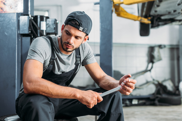 professional workman holding wrench and sitting in mechanic shop - Φωτογραφία, εικόνα