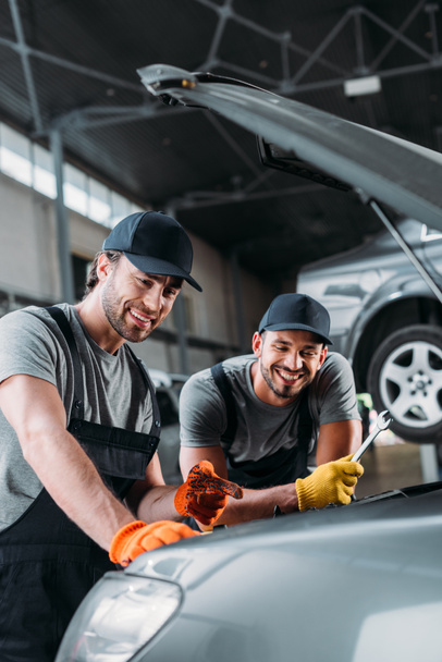 professional smiling auto mechanics repairing car in workshop - Photo, Image