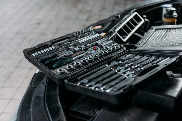 Werkzeugkoffer mit Schraubenschlüsseln und Metallmuttern liegt auf geöffneter Motorhaube in Werkstatt - Foto, Bild
