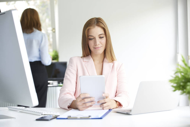 Attractive young businesswoman holding digital tablet in her hand and browsing on the internet while sitting at office desk and working. - Zdjęcie, obraz