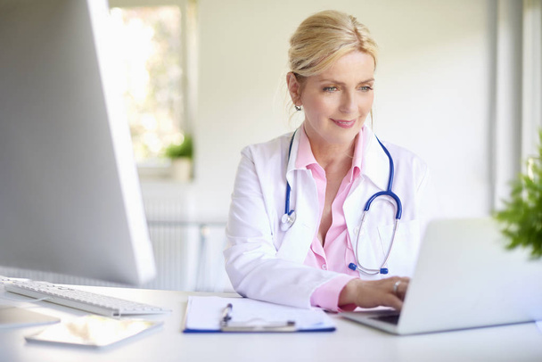 Mature femme médecin en utilisant son ordinateur portable tout en étant assis au bureau du médecin
. - Photo, image