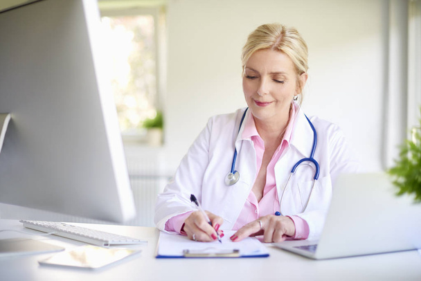 Zár-megjelöl szemcsésedik-ból mosolygó idős nő orvos használ a laptop, és írásban valami, miközben ül íróasztal, az orvosi rendelő.  - Fotó, kép