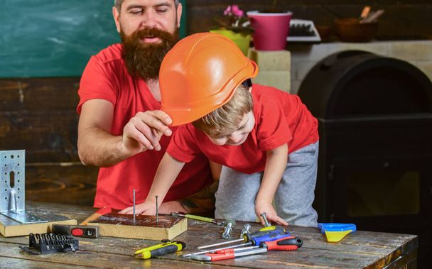 Отец, родители с бородой учат маленького сына использовать разные инструменты в школьной мастерской. Мальчик, ребенок занят игрой в защитный шлем, учится пользоваться инструментами с папой. Концепция отцовства
 - Фото, изображение