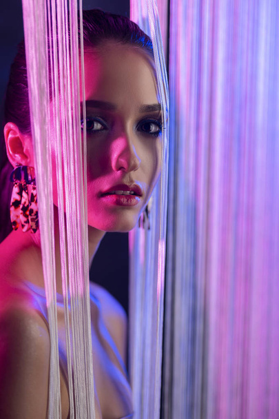 A beautiful glamorous brunette girl with naked shoulders wearing large elegant earrings peeps through a translucent curtain made of white threads. Multicolored artistic stage lighting. Copy space. - Fotografie, Obrázek
