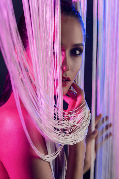 A beautiful glamorous brunette girl with naked shoulders wearing large elegant earrings peeps through a translucent curtain made of white threads. Multicolored artistic stage lighting. Copy space. - Foto, Bild