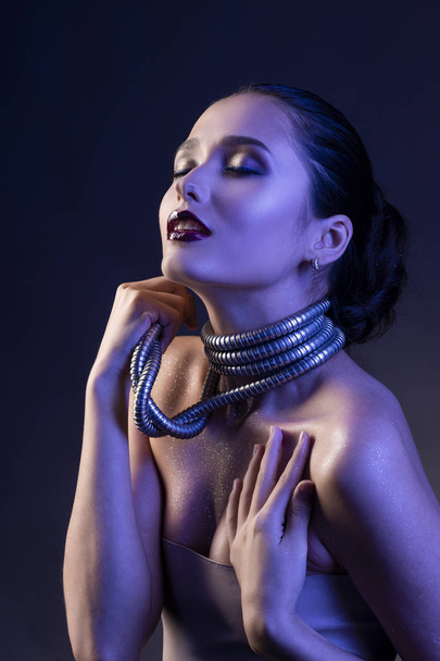 Beautiful brunette girl with naked shoulders and dark red lips make-up, wears a metal shower spiral hose wrapped around her neck. Concept and advertising low key photo illuminated in blue. Copy space - Фото, изображение