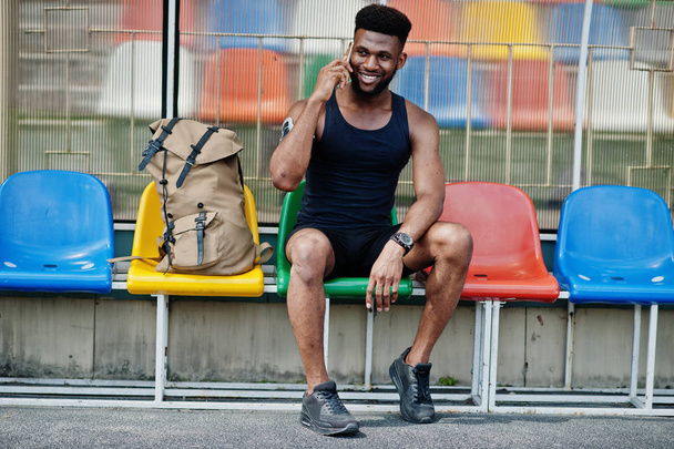 African american male athlete in sportswear with backpack sitting on chair at stadium and speaking on mobile phone. - Foto, Imagen