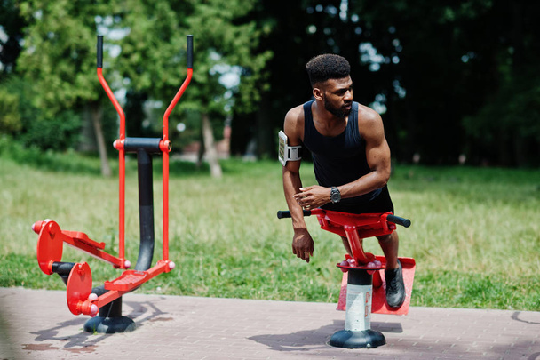 Hombre atleta afroamericano con funda de brazo deportivo para teléfono móvil, haciendo ejercicios en los simuladores de entrenamiento callejeros
. - Foto, Imagen