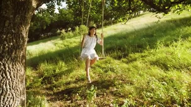 piękna dziewczyna z długimi włosami w białej sukni, kołysanie na huśtawce i uśmiechając się w parku lato latem. Zwolnionym tempie. - Materiał filmowy, wideo