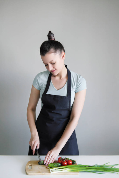 A woman in a kitchen apron cuts vegetables on a wooden board. - Φωτογραφία, εικόνα