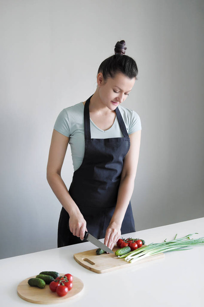 Una donna in un grembiule da cucina taglia le verdure su una tavola di legno
. - Foto, immagini