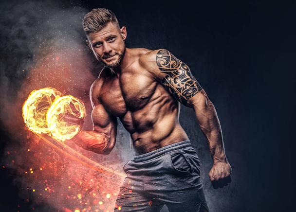 Powerful stylish bodybuilder with tattoo on his arm, doing the exercises on a biceps with dumbbell, looking at a camera. Fire art concept. - Photo, Image