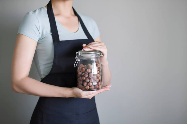 A woman in an apron holds a glass jar with hazelnuts in the kitchen in her hands. - Fotó, kép