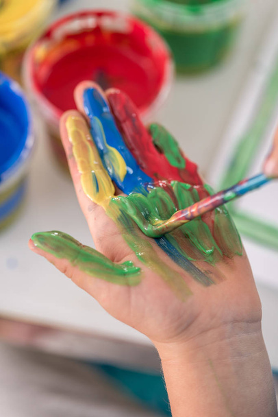 Creative little boy painting its hand with a paintbrush with colorful paint in a close up view of his palm. - Photo, image