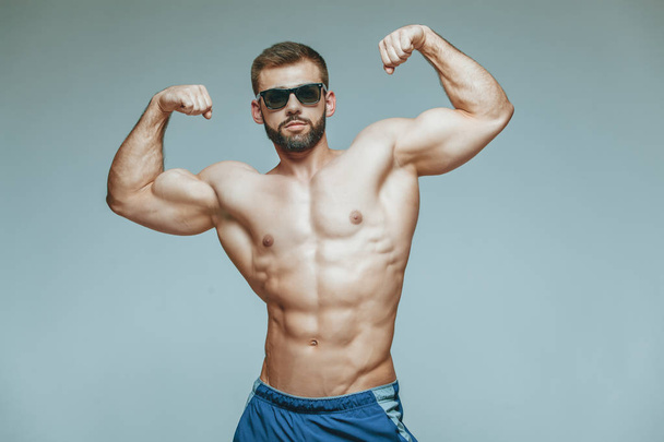 .bodybuilder posing. Beautiful sporty guy male power. Fitness muscled in blue shorts and sunglasses. on isolated grey background. Man with muscular torso. Strong Athletic Man Fitness Model Torso - Foto, Bild
