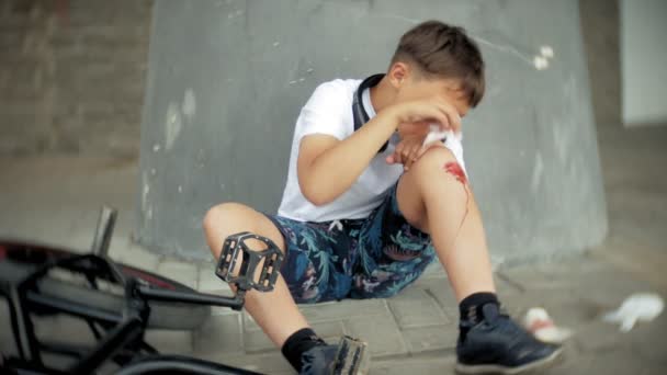 Un niño se sienta en un parque después de caer de una bicicleta, calma el dolor en la rodilla, es un peligroso paseo en bicicleta
 - Metraje, vídeo