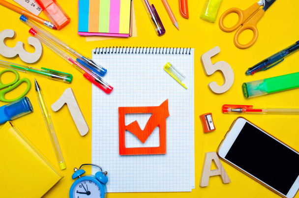 school elections concept. Election check box and school accessories on a desk on a yellow background. education. stationery, watches, colored pens, phone, markers. notebook. view from above. - Photo, Image