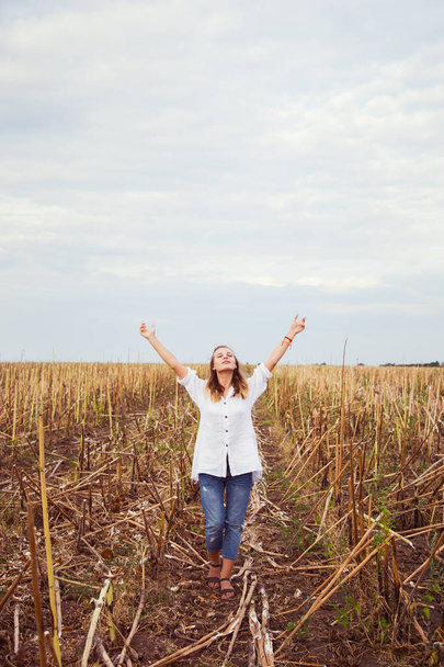 Young blond girl in casual clothes with arms raised enjoys her walking in field. Happy woman relaxing on countryside. Concept of freedom and wellness. - Photo, Image