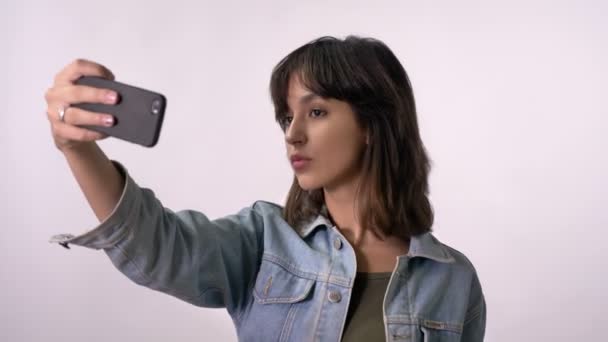 Young brunette girl is making selfie on smartphone, duck face, white background - Video, Çekim