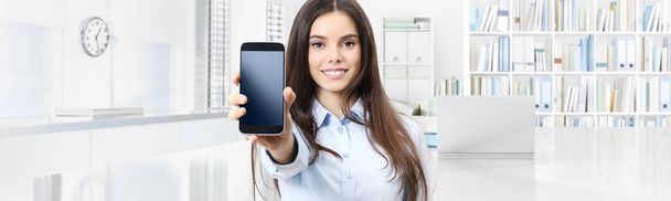 Улыбающаяся женщина показывает смартфон, изолированный на внутреннем фоне офиса
 - Фото, изображение