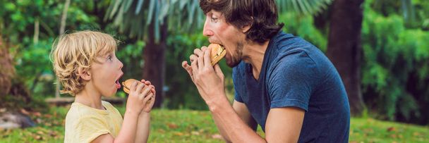 Portrait of a young father and his son enjoying a hamburgers in a park and smiling. BANNER long format - Photo, Image
