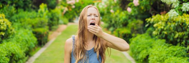 Young woman sneezes in the park against the background of a flowering tree. Allergy to pollen concept. BANNER long format - Foto, afbeelding