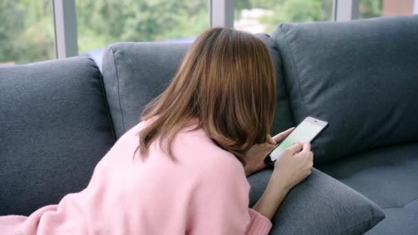 Mujer asiática joven usando el dispositivo de teléfono inteligente negro con pantalla verde. Mujer asiática sosteniendo el teléfono, páginas de desplazamiento mientras está sentado en el sofá en la sala de estar. Clave de croma
. - Metraje, vídeo