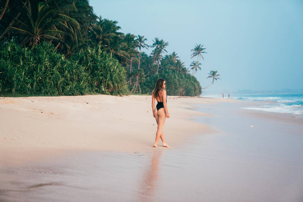 A slender young girl came to a deserted island, strolls along the white beach, admires the sea, wears a stylish bikini swimsuit, sunglasses. - 写真・画像