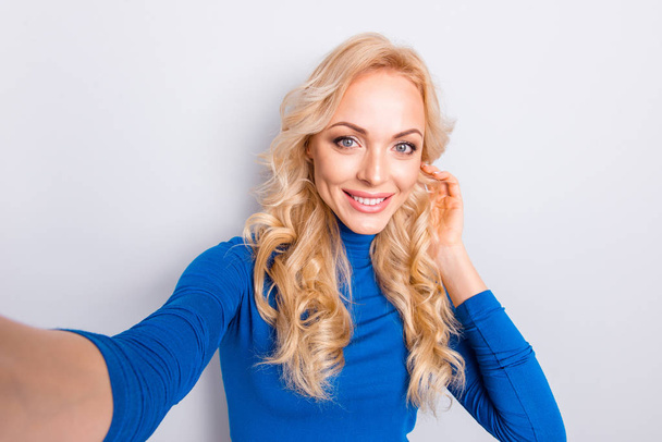 Self portrait of pretty, trendy, charming, lovely, nice, sexy, cute, blonde woman in blue turtleneck isolated on grey background shooting selfie on front camera holding hand on hair - Фото, зображення