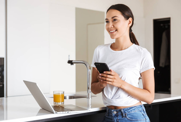 Smiling young asian woman using mobile phone while sitting on a kitchen with laptop computer and glass of orange juice - Zdjęcie, obraz