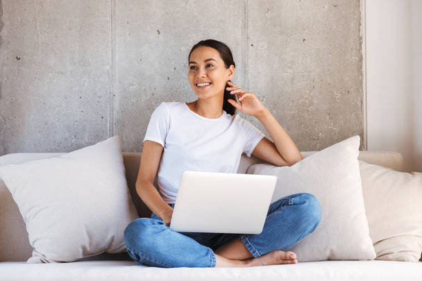 Pretty young asian woman using laptop computer while sitting on a couch at home - Photo, Image