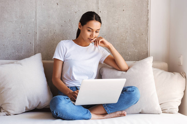 Pensive young asian woman using laptop computer while sitting on a couch at home - Photo, Image