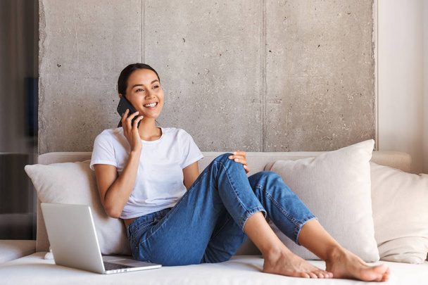 Smiling young asian woman talking on mobile phone while sitting on a couch at home with laptop computer - Photo, Image