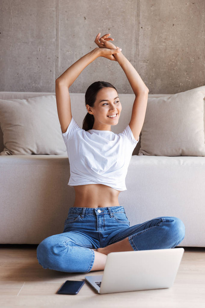 Happy young asian woman stretching her hands up while sitting at a couch at home with laptop computer - Foto, imagen