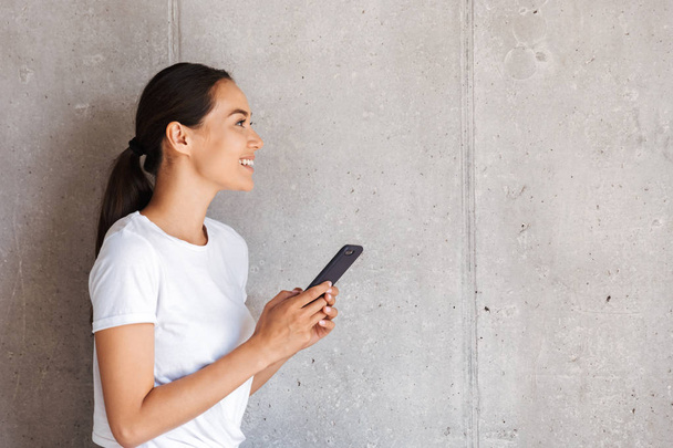 Sorridente giovane donna asiatica in possesso di telefono cellulare e guardando lontano a copiare spazio su sfondo grigio muro
 - Foto, immagini