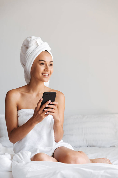 Cheerful young asian woman wrapped in a shower towel using mobile phone while sitting on bed - Фото, зображення