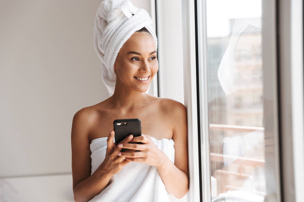 Smiling young asian woman wrapped in a shower towel using mobile phone while standing at the window - Foto, afbeelding
