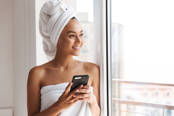 Cheerful young asian woman wrapped in a shower towel using mobile phone while standing at the window - Foto, imagen
