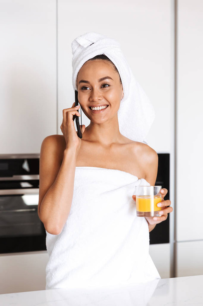 Smiling young asian woman wrapped in a shower towel talking on mobile phone while standing at the kitchen holding glass of juice - Foto, Imagem